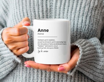 Può includere: Una tazza in ceramica bianca con un design nero. La tazza presenta la parola "Anne" in un carattere grande, seguita dal testo "[Fatma]" in un carattere più piccolo. Sotto il nome, c'è una definizione del nome in turco. La definizione recita: "Herkesin yerini alabilen, ancak hiç kimsenin yerini alamadigi kişi. Sizi dünyada en fazla sevebilecek insan, "fedakar" kelimesinin vücut bulmuş hali, koruyucu Melek Iyi ki varsın"