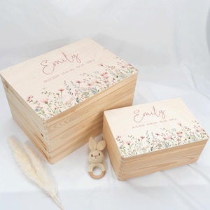 May include: Two wooden keepsake boxes with floral designs and the name "Emily" in pink script. The lids feature a delicate watercolor floral border. A small crochet bunny rattle is next to the boxes.