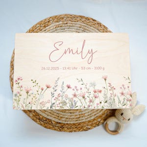May include: A light wooden plaque with the name "Emily" in pink script. The plaque features floral artwork along the bottom edge and includes the date, time, length (53 cm), and weight (3100 g).