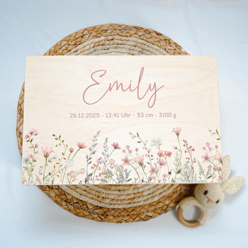 May include: A light wooden plaque with the name "Emily" in pink script. The plaque features floral artwork along the bottom edge and includes the date, time, length (53 cm), and weight (3100 g).