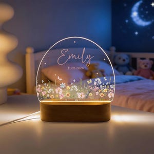 Könnte beinhalten: Personalisiertes Nachtlicht mit Holzsockel und klarer Acryloberfläche. Auf dem Acryl sind der Name "Emily" und das Datum "11.05.2026" sowie ein Blumendesign eingraviert. Das Licht strahlt warm.