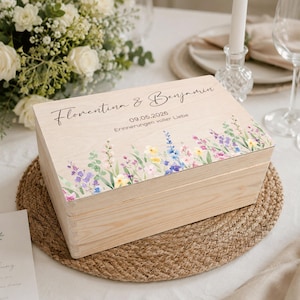 Könnte beinhalten: Eine hellfarbene Holzkiste mit floralem Design und Text. Die Kiste hat einen Aquarell-Rand aus bunten Wildblumen. Der Deckel ist mit den Namen "Florentina & Benjamin", dem Datum "09.05.2026" und dem Satz "Erinnerungen voller Liebe" beschriftet.