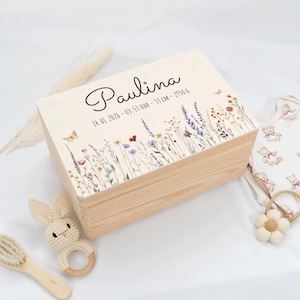 Könnte beinhalten: Eine hellfarbene Holzkiste mit dem Namen "Paulina" in Schreibschrift. Der Deckel ist mit einem Blumendesign und Schmetterlingen verziert. Auf der Box sind Datum, Uhrzeit, Länge (53 cm) und Gewicht (2950 g) aufgedruckt. Ein Babyrassel und eine Bürste liegen daneben.