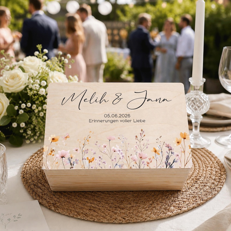 May include: A wooden box featuring the names "Melih & Jana" in script, with the date "05.06.2026". The box is adorned with a floral design of colorful wildflowers. It rests on a woven placemat, suggesting a celebratory occasion.