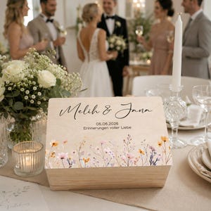 May include: A wooden keepsake box with a floral design and the names "Melih & Jana" in elegant script. The box features the date "05.06.2026" and the phrase "Erinnerungen voller Liebe". The box is on a table with wedding decorations.