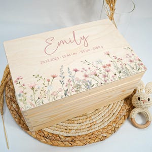 May include: A light-colored wooden keepsake box with a floral design and the name "Emily" in pink script. The box features a border of pink and white flowers and the date, time, length, and weight of the baby. A crocheted bunny rattle is next to the box.