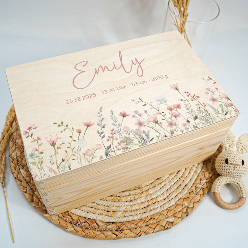 May include: A light-colored wooden keepsake box with a floral design and the name "Emily" in pink script. The box features a border of pink and white flowers and the date, time, length, and weight of the baby. A crocheted bunny rattle is next to the box.