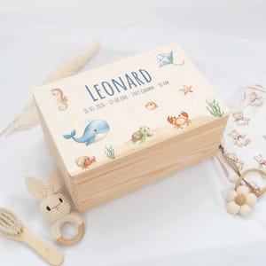 Puede incluir: Una caja de recuerdos de madera de color claro con un tema marino. La caja está decorada con ilustraciones de criaturas marinas, incluyendo una ballena, un cangrejo y un caballito de mar. El nombre "LEONARD" está impreso en la tapa, junto con los detalles del nacimiento.