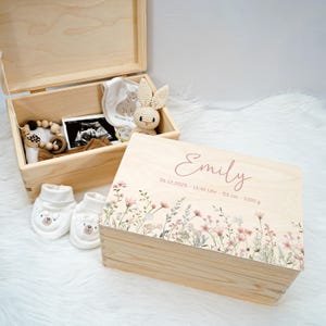 May include: A wooden keepsake box with a floral design and the name "Emily" printed on the lid. The box contains baby items, including a bib, booties, a rattle, and a stuffed bunny. The lid also has the baby's birth details.