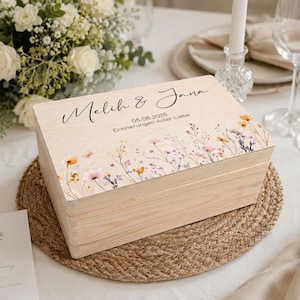 Könnte beinhalten: Eine hellfarbene Holzkiste mit floralem Design und den Namen "Melih & Jana" auf dem Deckel. Die Kiste trägt auch das Datum "05.06.2026" und den Satz "Erinnerungen voller Liebe". Die Kiste steht auf einer gewebten Matte.