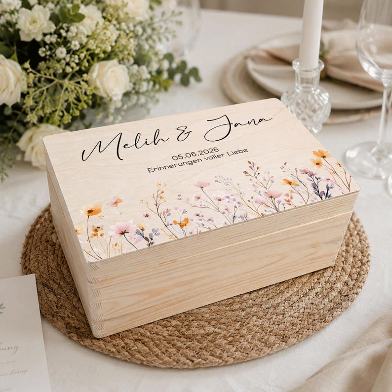 May include: A light-coloured wooden box with a floral design and the names "Melih & Jana" printed on the lid. The box also has the date "05.06.2026" and the phrase "Erinnerungen voller Liebe" printed on the lid. The box is sitting on a woven mat.
