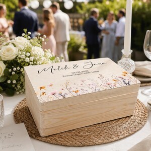 May include: A wooden box with a floral design and text. The box is rectangular and sits on a woven placemat. The lid features the names "Melih & Jana" and the date 05.06.2026. The box is light-colored.