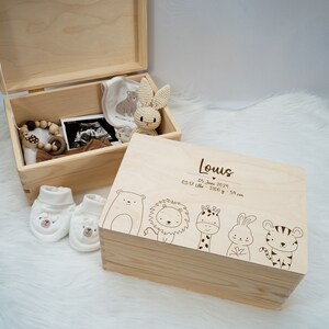 Op de afbeelding: Een houten herinneringsdoos met een lichte houtkleur. De deksel is gegraveerd met de naam "Louis", de datum "05 juni 2024", de tijd "03:17 Uhr", het gewicht "3100 g" en de lengte "54 cm". De deksel heeft ook een rij cartoondieren, waaronder een beer, een leeuw, een giraffe, een konijn en een tijger. De doos is open en bevat verschillende babyartikelen, waaronder een paar witte slofjes, een houten rammelaar, een speen, een slabber en een gehaakt konijn.