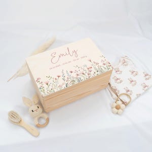 May include: A light-colored wooden keepsake box with floral accents and the name "Emily" in cursive. The box is surrounded by baby items, including a bunny rattle, a brush, and a onesie.