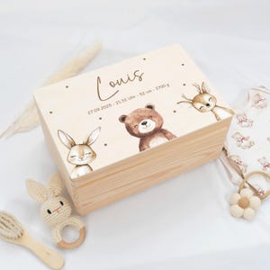 Puede incluir: Una caja de madera clara decorada con ilustraciones de un conejito, un oso y un ciervo. La caja está personalizada con el nombre "Louis" y detalles de nacimiento. Un sonajero de conejo de ganchillo y ropa de bebé están cerca.