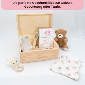 May include: A wooden box with baby items, including a teddy bear, a bunny rattle, a baby brush, a card with a pink elephant, and a baby onesie with teddy bear patterns. The text at the top says "The perfect gift idea for birth, birthday or baptism."