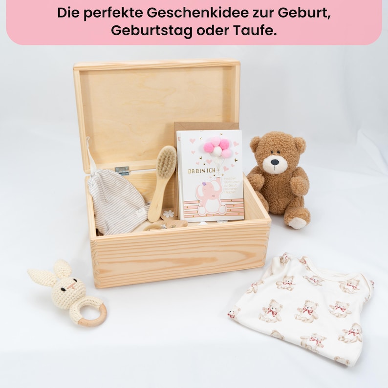 May include: A wooden box with baby items, including a teddy bear, a bunny rattle, a baby brush, a card with a pink elephant, and a baby onesie with teddy bear patterns. The text at the top says "The perfect gift idea for birth, birthday or baptism."