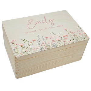May include: A wooden keepsake box with a light-colored finish. The lid features the name "Emily" in pink script, along with birth details. Delicate floral artwork in pink, white, and green adorns the lid's edge. The box is rectangular with dovetail joints.
