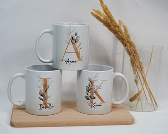 Può includere: Tre tazze in ceramica bianca con lettere dorate e disegni floreali. Le tazze sono impilate l'una sull'altra. La tazza in alto ha la lettera "A" e il nome "Anna" stampato su di essa. La tazza nel mezzo ha la lettera "I" e il nome "Isabella" stampato su di essa. La tazza in basso ha la lettera "K" e il nome "Karin" stampato su di essa.