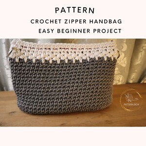 May include: A handmade crochet handbag in grey and white. The bag has a zip closure and metal ring accents. The text "PATTERN CROCHET ZIPPER HANDBAG EASY BEGINNER PROJECT" is displayed at the top of the image.