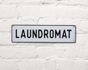 May include: White metal sign with black lettering that reads "LAUNDROMAT".