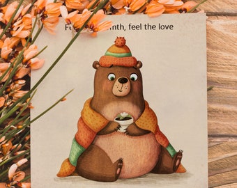 Puede incluir: Un oso marrón con una bufanda verde y naranja y un gorro rojo y naranja está sosteniendo una taza de té. El texto "For the month, feel the love" está escrito encima del oso.