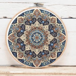 Op de afbeelding: Een ronde borduurring met een gedetailleerd, patroonontwerp. Het ontwerp bevat tinten blauw, bruin en crème, waardoor een mandala-achtig patroon ontstaat. De ring is gemaakt van lichtgekleurd hout en de stof is strak in het frame gespannen.
