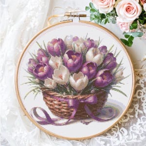 May include: Cross-stitch embroidery of a floral bouquet in a basket. The flowers are purple and white, with green leaves. The basket is brown with a purple ribbon. The embroidery is set in a wooden hoop.