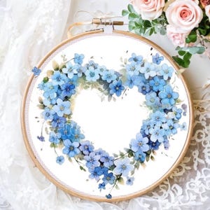 May include: A round embroidery hoop with a white background and a heart-shaped wreath of blue forget-me-not flowers.