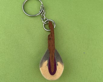 May include: A handmade felt keychain in the shape of a musical instrument. The keychain features a teardrop-shaped body with a light brown wooden neck and a purple accent. The keychain is attached to a silver metal ring.