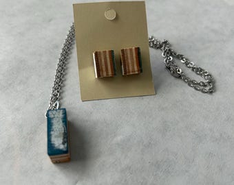 May include: A jewelry set featuring a necklace and matching earrings. The necklace has a rectangular pendant with blue and white accents, suspended from a silver chain. The earrings are square studs with a striped pattern in shades of brown, green, and white.