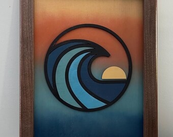 May include: A framed wall art piece featuring a layered wooden cutout of a stylized ocean wave and a sun inside a circular black border. The background displays a gradient transition from orange at the top to deep blue at the bottom.