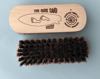 May include: A wooden brush with dark bristles. The brush handle is light-colored and features the text "THE QUIK SAND BRUSH" with a surfboard graphic and footprints. The brush is made by Second Beat Studios LLC.