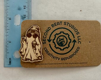 May include: A laser-cut wooden ghost pin wearing a beanie and sunglasses while holding a coffee cup and making a peace sign. The pin is attached to a brown card with the text Second Beat Studios LLC Creativity Repurposed. A blue ruler shows the pin is approximately 2.5 inches tall.