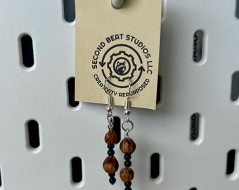 May include: Handmade earrings with a unique design. Each earring features a combination of brown, textured beads and small black beads, suspended from silver-colored hooks. The earrings are displayed with a tag that reads "SECOND BEAT STUDIOS LLC CREATIVITY REPURPOSED."