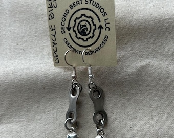 May include: Upcycled bike chain earrings with small silver dinosaur charms. The earrings are attached to a tag that reads "UPCYCLE BIKE CHAINS" and "SECOND BEAT STUDIOS LLC CREATIVITY REPURPOSED".