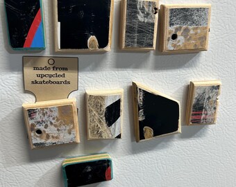 May include: A collection of upcycled skateboard magnets in various shapes and sizes. The magnets feature black, red, blue, and white paint designs. The text "made from upcycled skateboards" is also visible.