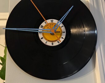 May include: A black vinyl record clock with orange and blue clock hands. The clock face features a yellow and white label with black text and numbers. The clock is mounted on a white wall.