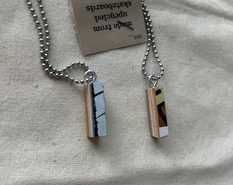 May include: Two silver ball chain necklaces each with a small rectangular pendant. The pendants are made from upcycled skateboards and feature colorful designs. A tag reads "made from upcycled skateboards."