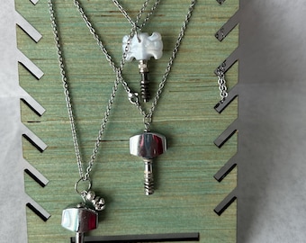 May include: Three silver-toned necklaces with pendant charms displayed on a green wooden stand. The charms include a hammer, a decorative white charm, and a hammer with two small ball charms. The necklaces are on a light green wooden display.