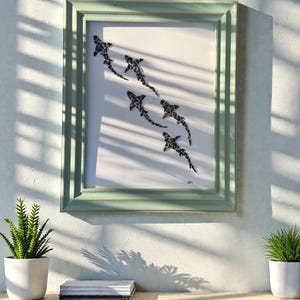 May include: A framed art print featuring a school of sharks in black and white. The artwork is set against a white background and framed in a light green wooden frame. The print is displayed on a wooden table with two small potted plants.