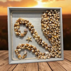 May include: A square, white-framed seashell art piece. The artwork features a design made of small, tan seashells, a starfish, and a rope border. The background is a light wood color.