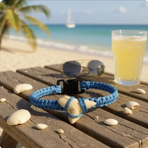 May include: A blue braided bracelet with a shark tooth pendant, resting on a wooden surface with seashells, sunglasses, and a glass of beer. The background shows a beach with a palm tree and ocean.
