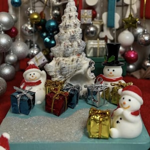 May include: A Christmas-themed display featuring a large seashell, miniature snowmen, and wrapped gift boxes. The snowmen wear red hats and scarves. The gifts are in blue, red, silver, and gold wrapping. Ornaments hang in the background.