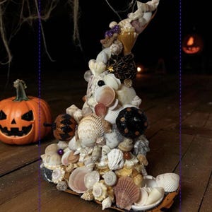 May include: A whimsical Halloween witch hat decorated with various seashells, small black decorative balls, and purple beads. An orange jack-o'-lantern and a pumpkin are in the background, set on a wooden surface.