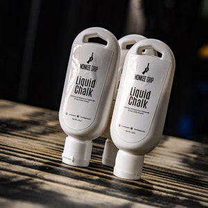 May include: Two white bottles of liquid chalk with black text that reads "MONKEE GRIP Liquid Chalk 100% Natural Magnesium Carbonate & Food Grade Ethanol Net Wt 5.2 fl oz (155ml)"