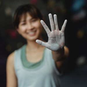 May include: A woman with chalk on her hand, smiling at the camera. She is wearing a light grey tank top and is in a climbing gym.