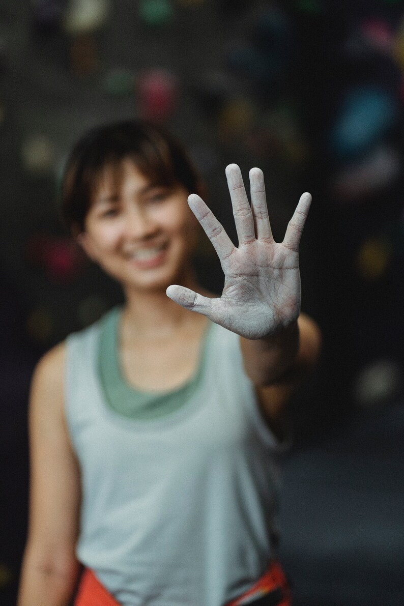 May include: A woman with chalk on her hand, smiling at the camera. She is wearing a light grey tank top and is in a climbing gym.