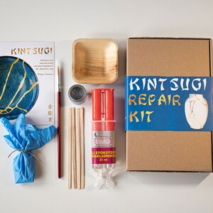 Puede incluir: Kit de reparación Kintsugi con un tazón azul y dorado, un tazón pequeño de madera, un frasco de polvo dorado, un tubo de epoxi, dos palitos de madera, un par de guantes azules, un pincel y una caja marrón con el texto "Kintsugi Repair Kit" y una imagen de un jarrón con grietas doradas.