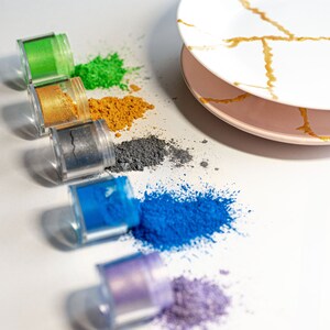 May include: Six small jars of colorful powder pigments, arranged in a row from green to purple, with a small amount of each powder spilled out in front of the jar.